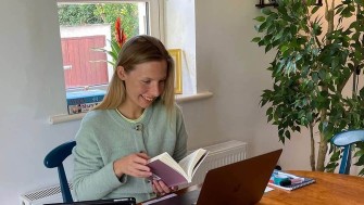 A woman flips through a book whilst seated in front of her open laptop