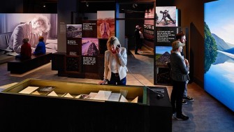 A photo of the Seamus Heaney: Listen Now Again exhibition space with visitors examining the manuscripts on display, watching a film and walking around the space.