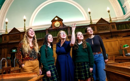 Stiúrthóir Gníomhach NLI Katherine McSharry agus ceathrar ban óg ina timpeall, agus í ag comharthú lena lámh i bPríomhsheomra Léitheoireachta NLI