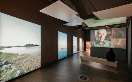 Section of 'Seamus Heaney: Listen Now Again' exhibition space. To the left is a row of scenic images of bodies of water. To the right a visitor sits on a bench watching a video of Seamus Heaney speaking.