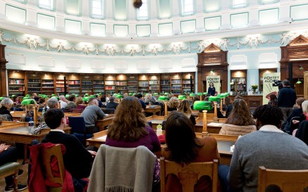 Suíonn cuairteoirí sa Seomra Léitheoireachta i Leabharlann Náisiúnta na hÉireann, ag amharc ar bhean ag labhairt le linn imeacht Poetry Aloud 