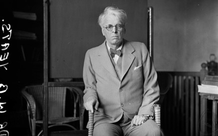 WB Yeats ag caitheamh culaithe, ina shuí i gcathaoir agus ag baint beagáinín taca as bata siúil 