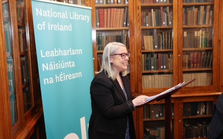 Bean ag labhairt d’ardán agus seilfeanna leabhar sa chúlra