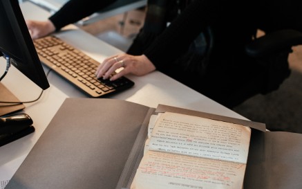 Image of hands typing at a computer with manuscript on desk